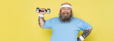 positive plus size man in blue t-shirt training with dumbbell isolated on yellow, banner