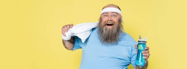 excited chubby man with sports bottle and towel smiling at camera isolated on yellow, banner