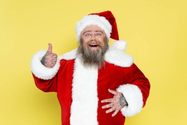 happy plus size santa claus showing thumb up and touching tummy isolated on yellow