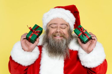 pleased and chubby santa claus with closed eyes holding gift boxes isolated on yellow