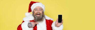 cheerful plus size santa claus pointing at mobile phone with blank screen isolated on yellow, banner