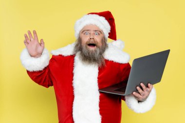 astonished overweight santa claus with laptop waving hand isolated on yellow