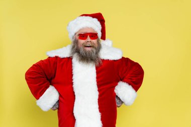 cheerful overweight santa claus in red sunglasses standing akimbo isolated on yellow