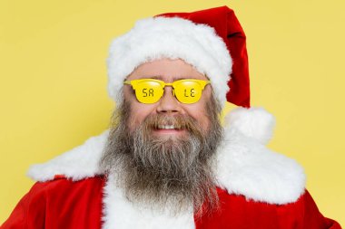 smiling and chubby man in christmas costume and glasses with sale lettering isolated on yellow