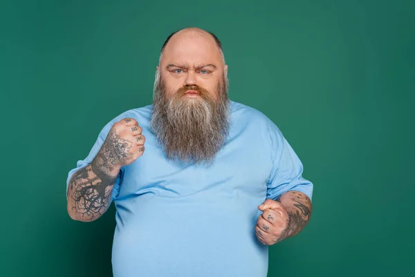 angry overweight man with tattoos and beard showing clenched fists isolated on green