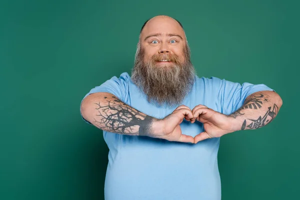 happy plus size man with beard and tattoos showing heart sign isolated on green