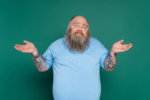 confused plus size man with beard and tattoos showing shrug gesture isolated on green