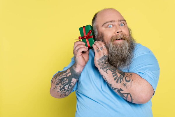 curious overweight man with beard holding green gift box isolated on yellow