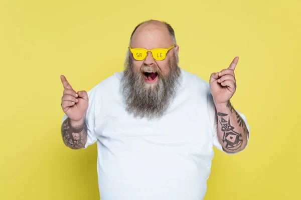 bold and bearded overweight man in glasses with sale lettering pointing with fingers isolated on yellow