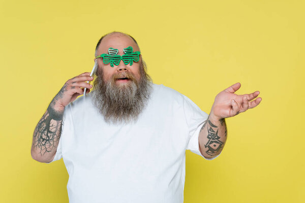 бородатый толстяк в очках в форме клевера разговаривает на смартфоне, изолированном на желтом