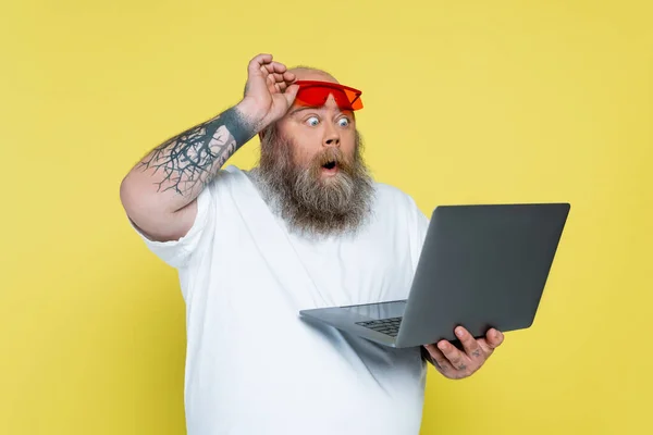surprised plus size man holding red sunglasses while looking at laptop isolated on yellow
