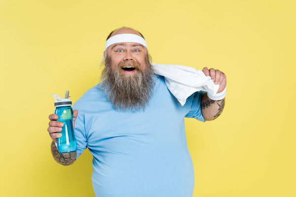 cheerful plus size man with sports bottle and towel looking at camera isolated on yellow