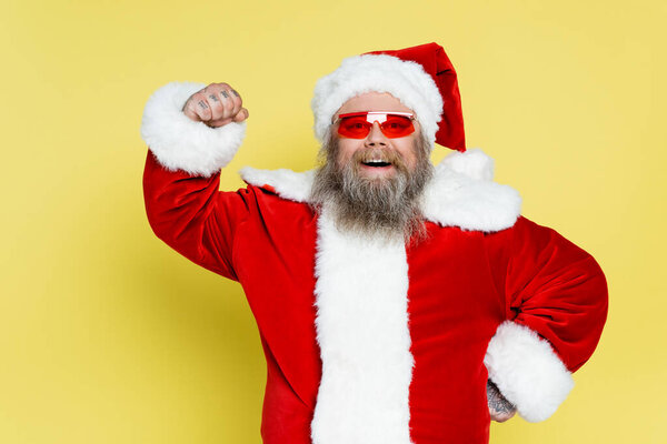 happy plus size santa claus in trendy sunglasses demonstrating power isolated on yellow