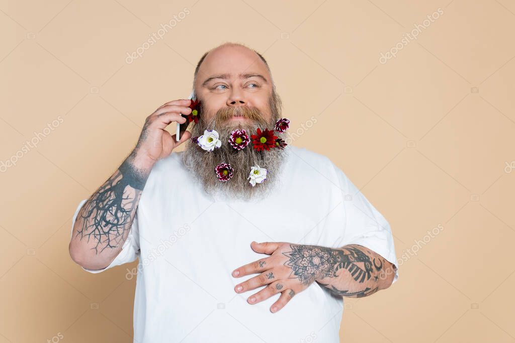 joyful man with flowers in beard talking on mobile phone isolated on beige