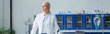Blonde scientist in white coat standing near flask with liquid in lab, banner 
