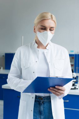 Beyaz önlüklü ve koruyucu maskeli bir bilim adamı laboratuarda çalışırken elinde bir pano tutuyor.