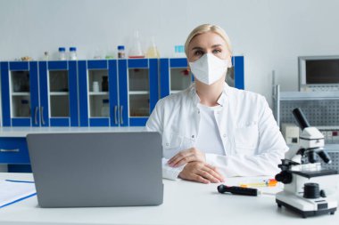 Koruyucu maskeli bilim adamı laboratuvarda mikroskop ve dizüstü bilgisayara bakıyor. 