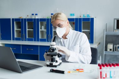 Scientist in protective mask using microscope near test tubes in laboratory 