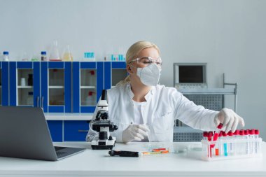Scientist in protective mask taking test tube near laptop and microscope in lab 