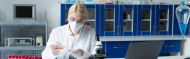 Scientist in protective goggles and mask holding petri dish and test tube in lab, banner 