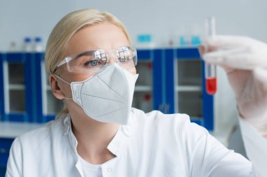 Gözlüklü ve koruyucu maskeli sarışın bilim adamı laboratuarda bulanık test tüpü tutuyor. 