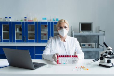 Scientist in protective mask looking at camera near test tubes and microscope in lab  clipart