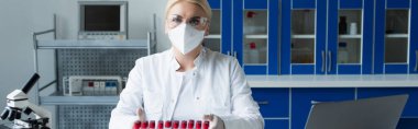 Scientist in goggles looking at camera near test tubes and microscope in lab, banner 