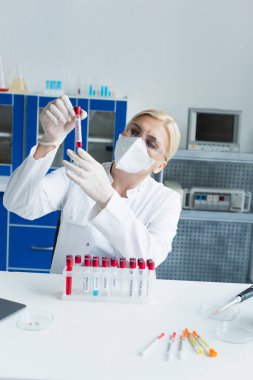 Gözlüklü ve maskeli bilim adamı test tüpü tutuyor. Laboratuvarda şırıngaların yanında maymun çiçeği harfleri yazıyor. 