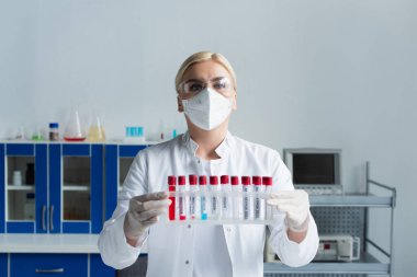 Gözlüklü ve beyaz önlüklü bir bilim adamı laboratuarda maymun çiçeği harfli test tüpleri tutuyor. 