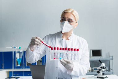 Koruyucu maskeli bir araştırmacı laboratuarda maymun çiçeği harfleriyle test tüpleri tutuyor. 