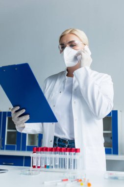 Koruyucu maskeli bilim adamı akıllı telefondan konuşuyor ve laboratuarda test tüplerinin yanında pano tutuyor. 