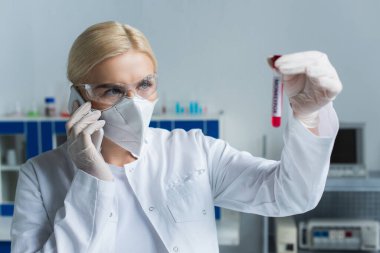 Gözlüklü bilim adamı akıllı telefondan konuşuyor ve laboratuardaki test tüplerine bakıyor. 