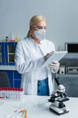 Laboratuvarda test tüplerinin yanında dijital tablet ve mikroskop kullanan koruyucu maskeli bilim adamları 