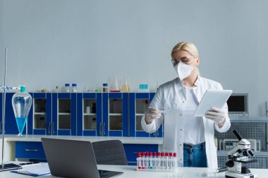 Koruyucu maskeli bilim adamı laboratuarda petri kabı ve dijital tablet tutuyor.