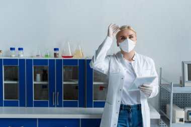 Koruyucu maskeli bilim adamı gözlük ve dijital tableti laboratuvarda tutuyor. 