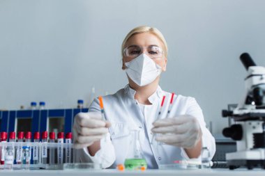 Mikroskop yanında bulanık şırıngalar ve laboratuvar tüpleri tutan maskeli bilim adamının düşük açılı görüntüsü 