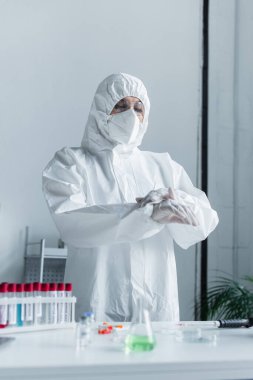 Scientist in protective suit and mask standing near blurred test tubes in lab 