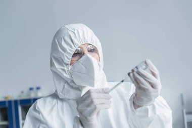 Koruyucu kıyafetli ve gözlüklü bilim adamı laboratuarda şırınga ve aşı tutuyor. 