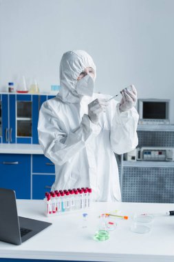 Tehlikeli madde giysisi giyen bilim adamı laboratuarda test tüplerinin yanında şırınga ve aşı tutuyor. 