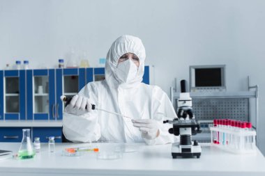 Scientist in hazmat suit working with pipette and glass near test tubes in lab 