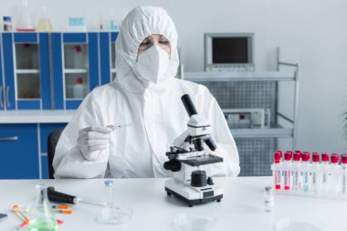 Scientist in hazmat suit holding glass near microscope and test tubes in lab 