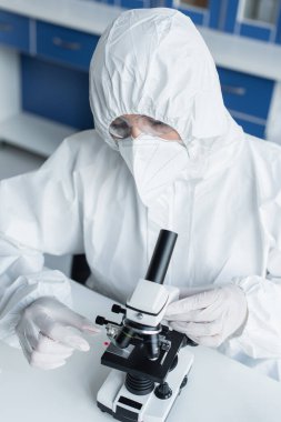 Scientist in hazmat suit holding glass near microscope in lab 