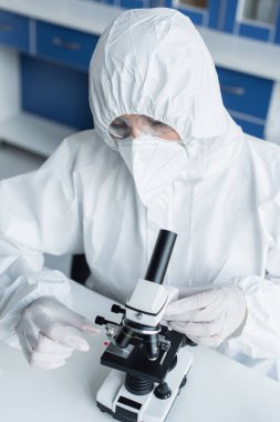 Laboratuvarda mikroskop yanında cam tutan tehlikeli madde giysisi giyen bilim adamı. 