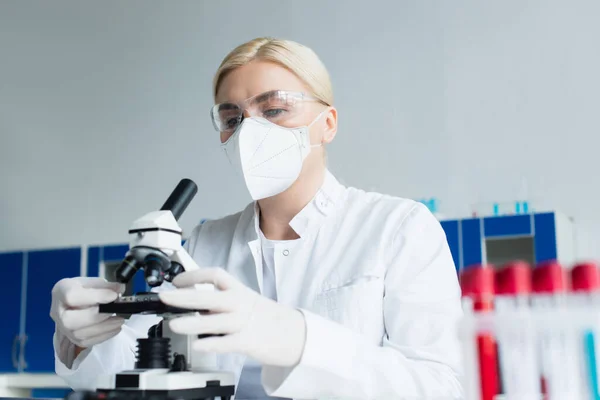 Scientist in goggles and latex gloves using microscope near blurred test tubes in lab