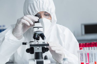 Scientist in hazmat suit using microscope near test tubes in lab 