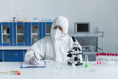Tehlikeli madde giysisi giyen bilim adamları mikroskop ve laboratuvar tüplerinin yanında panoya yazı yazıyorlar. 