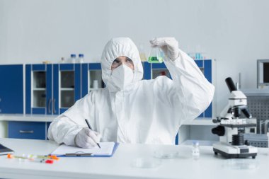 Tehlikeli madde giysisi giyen bilim adamı laboratuardaki panoda matara ve yazı işleriyle uğraşıyor.
