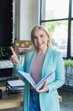 Smiling psychologist holding paper folder and pointing with finger in consultation room 