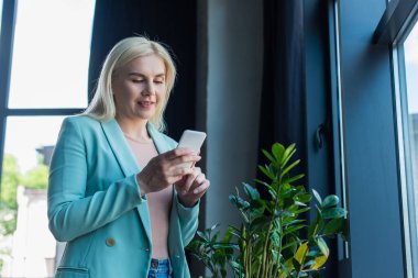 Gülümseyen psikolog danışma odasındaki bitkilerin yanında akıllı telefon kullanıyor. 