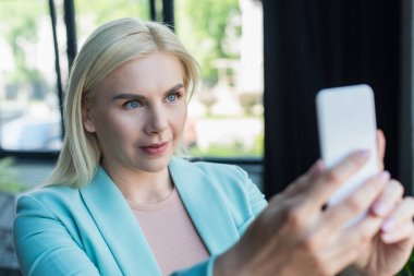 Blonde psychologist having video call on blurred smartphone in consultation room 